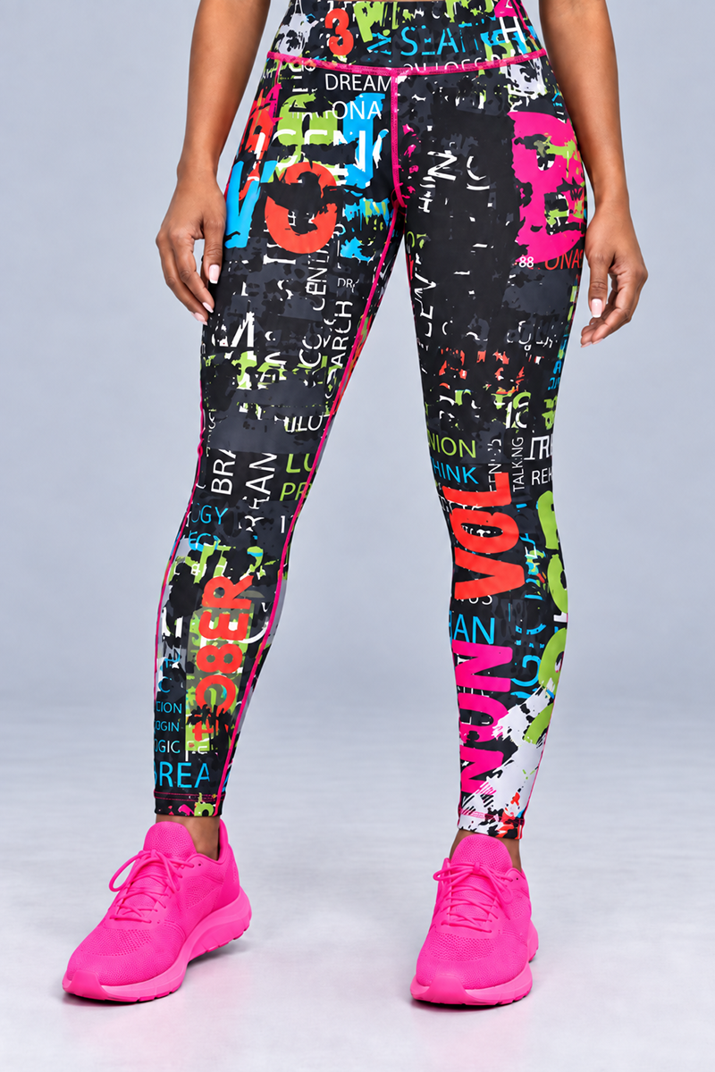 A person stands facing forward, wearing the Electric Letters Tights in Multi, which feature a vibrant graphic print with colorful words and designs. They are paired with bright pink athletic shoes. The background is plain light gray, and only the lower torso and legs are visible.