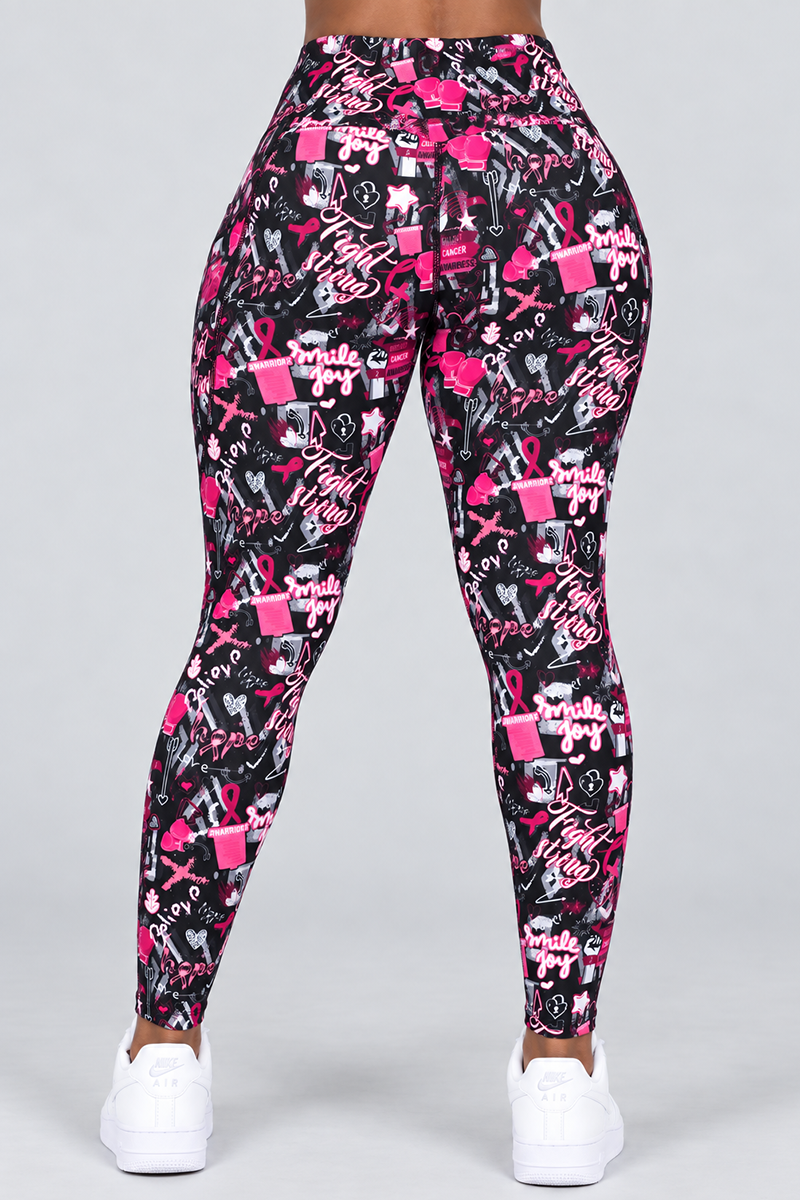 A person is shown from the waist down wearing the Fight Strong Tights—women’s workout leggings crafted from a polyester-spandex blend with black fabric decorated in pink and white breast cancer awareness motifs and supportive text. The comfortable tights are paired with white sneakers as the individual stands against a plain gray background.