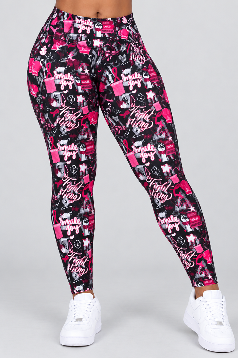 A person is wearing Fight Strong Tights—high-waisted women’s workout leggings in a multi-color breast cancer awareness design with ribbons, hearts, and inspiring words. Made from a polyester-spandex blend, these tights offer comfortable 4-way stretch and flexibility. They are paired with white sneakers against a plain gray background.