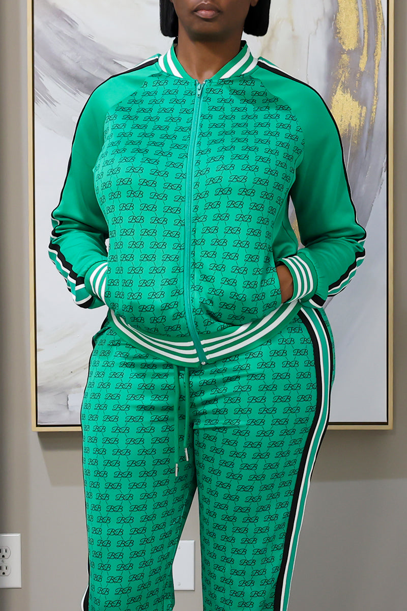 Wearing the Monogram Track Jacket, a relaxed fit green tracksuit with a repeating black motif and white-striped details, this individual stands hands-in-pockets before an abstract painting. The stretchy fabric offers both comfort and style.