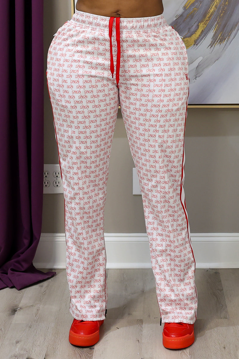 A person wearing BB Monogram Sweatpants featuring a relaxed fit and a red logo pattern, with red drawstrings and red sneakers, stands indoors on a wooden floor with a purple curtain and abstract wall art behind them.