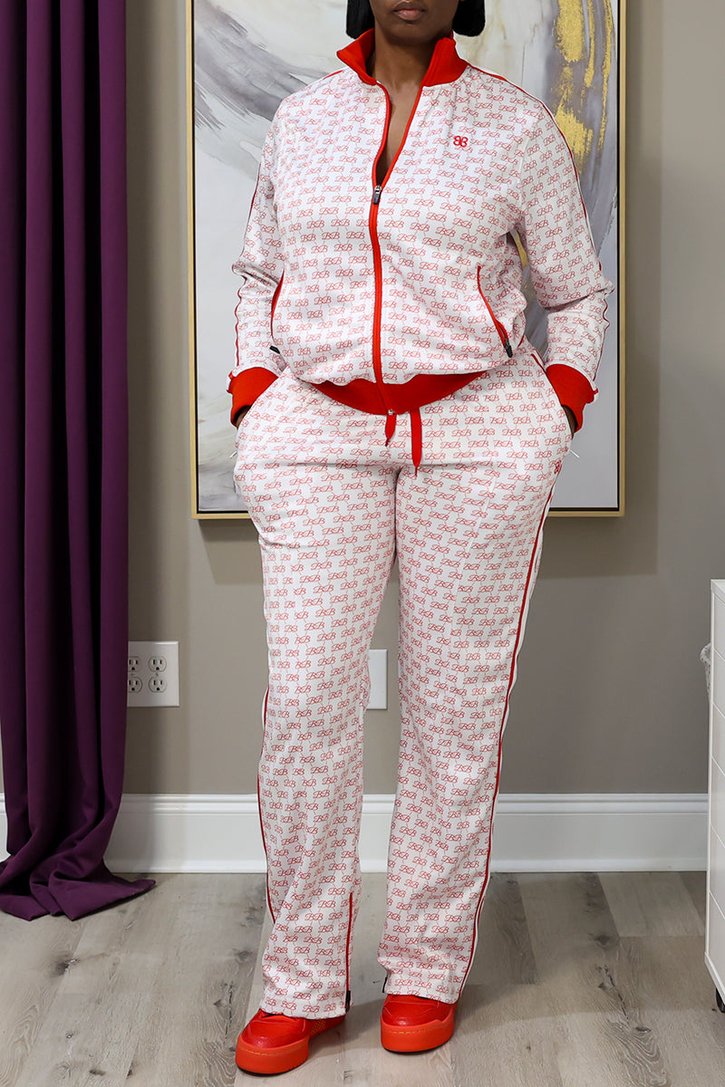 A person wears BB Monogram Sweatpants, paired with a matching white and red tracksuit and red shoes, standing indoors with hands in pockets. A painting and part of a purple curtain are visible in the background.