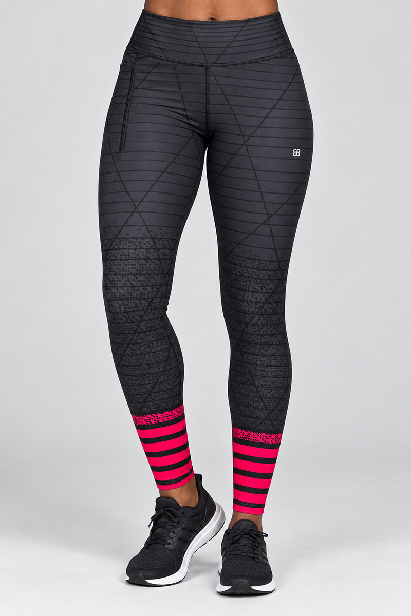 A person is standing against a plain, light gray background, wearing the Epic Tights in Black-Pink—athletic leggings crafted from a polyester spandex blend. These tights feature geometric patterns and red-striped accents near the ankles, offering a body-contouring fit for both style and performance. The look is paired with black athletic shoes.