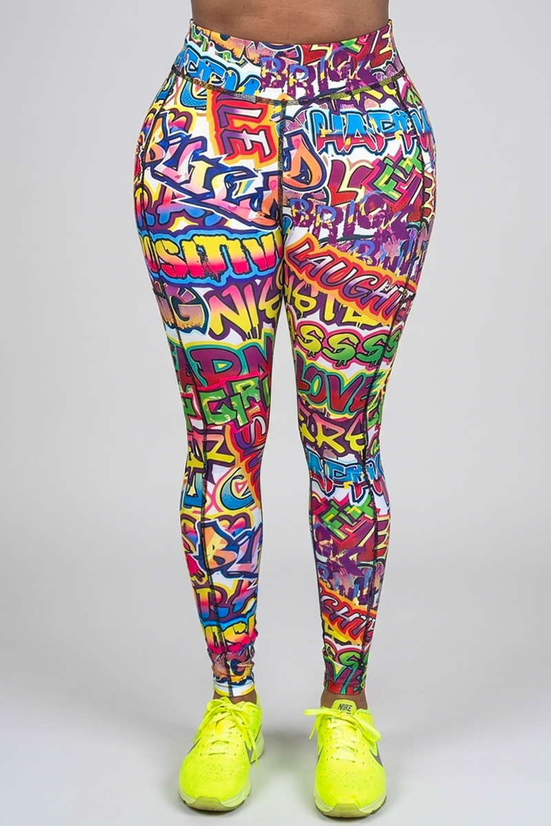 Model wearing Graffiti Tights in multi, featuring vibrant graffiti-inspired patterns and words, paired with bright yellow athletic shoes, standing against a plain gray background.