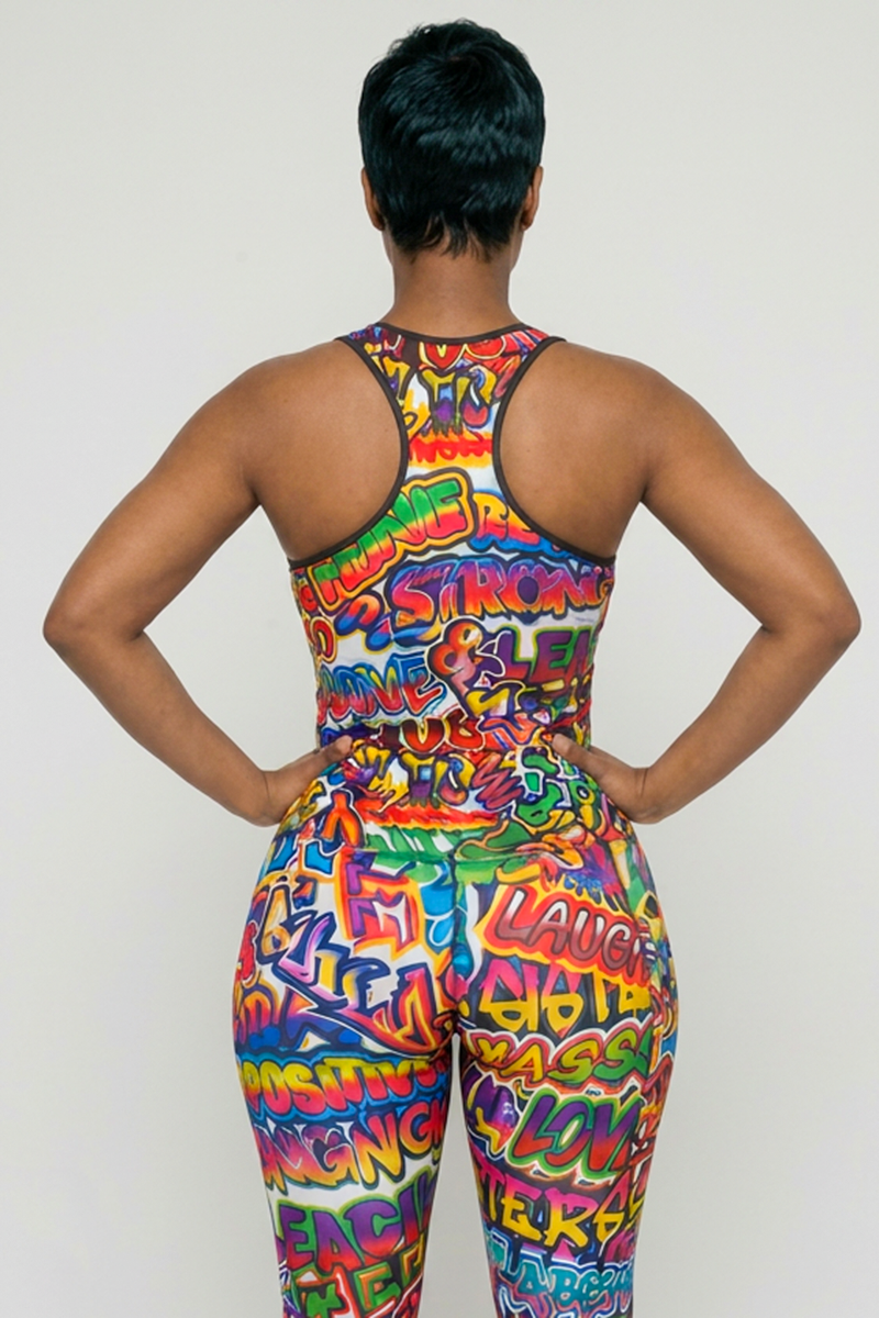 A person stands with hands on hips, wearing the Graffiti Tank Top—a colorful, graffiti-print polyester spandex top with 4-way stretch and moisture-wicking features. Words like “STRONG,” “LAUGH,” and “LOVE” stand out on a bright background.