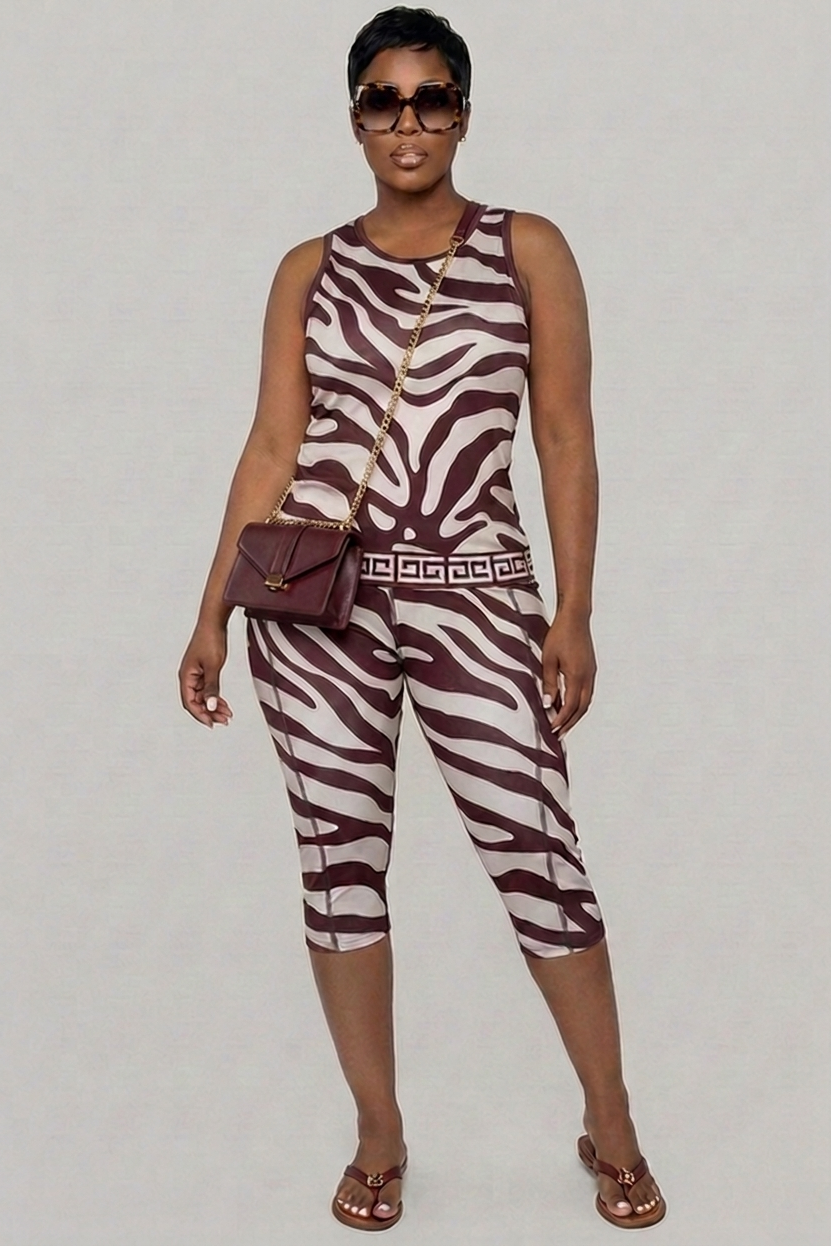 A woman poses against a plain background in ZéLuxe Capris with a bold zebra print, paired with a sleeveless top, short hair, sunglasses, sandals, and a burgundy crossbody purse featuring a gold chain strap. The stretchy ZéLuxe Capris provide 4-way comfort.