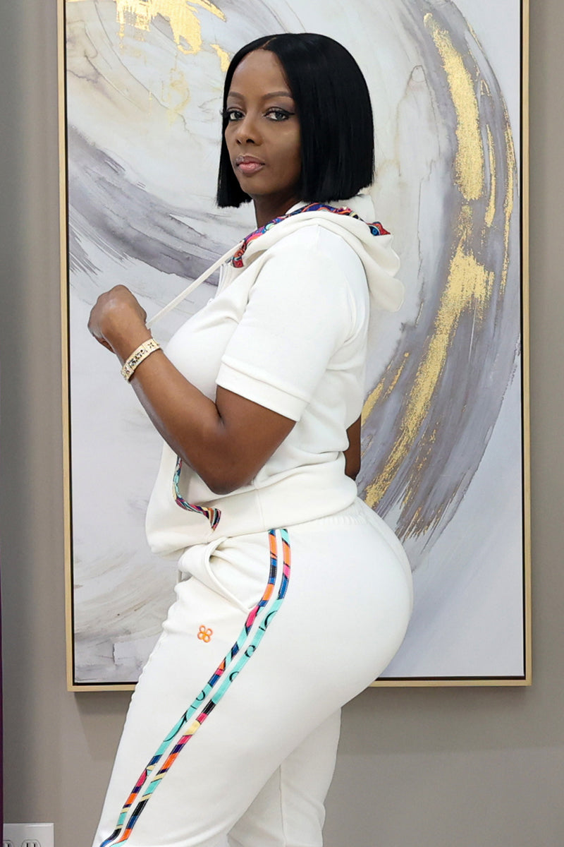 A woman stands before an abstract painting, wearing the Jet Set Short Sleeve Hoodie—white with kangaroo pockets and colorful trim—paired with matching pants, and glances over her shoulder at the camera.
