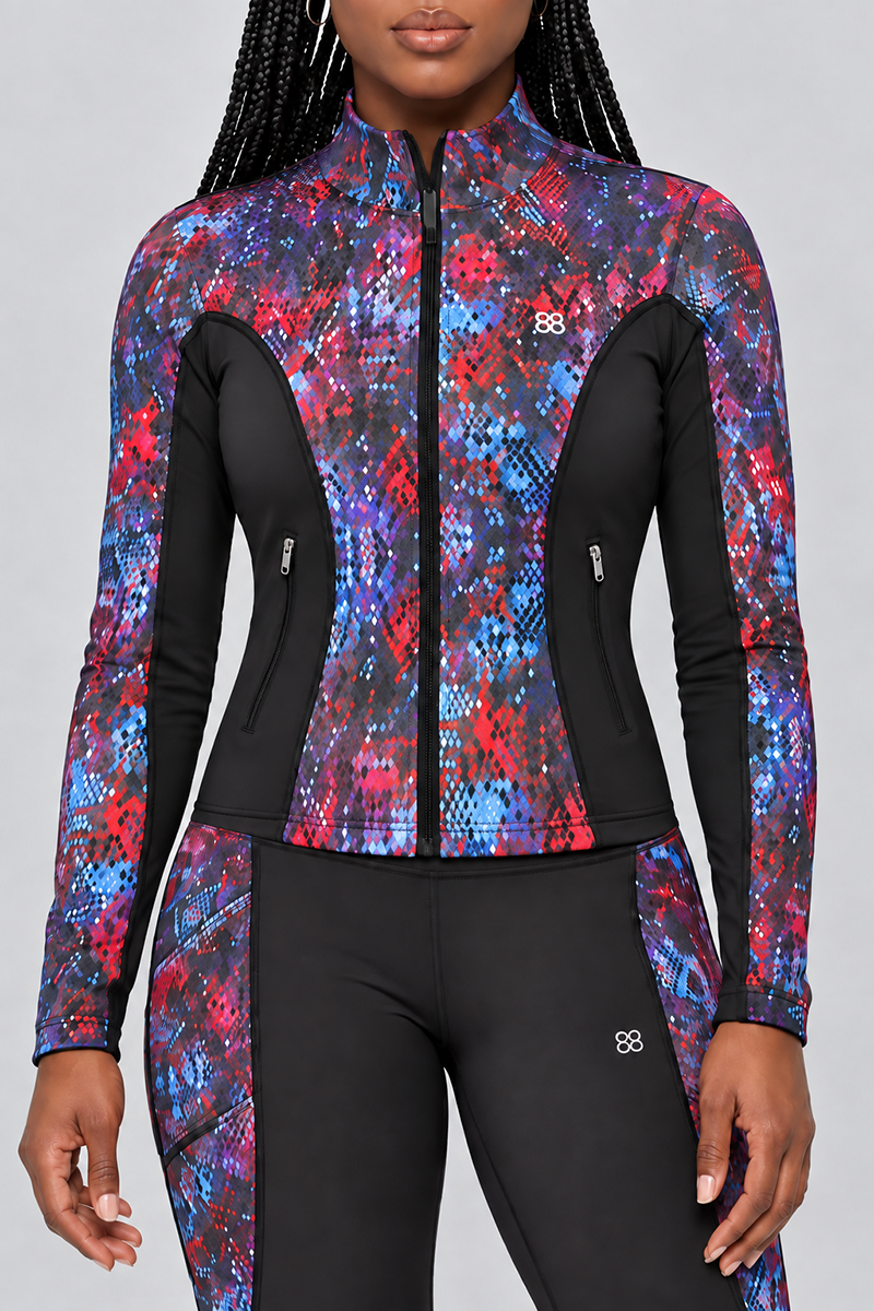 A woman wearing the Kario Jacket—a fitted, zip-up athletic piece paired with matching leggings—showcases a vibrant red, blue, and purple abstract pattern accented by black panels. Both the jacket and leggings offer a body-contouring fit crafted from soft polyester-spandex fabric with 4-way stretch for ultimate comfort and flexibility. Small white logos complete the sleek look.