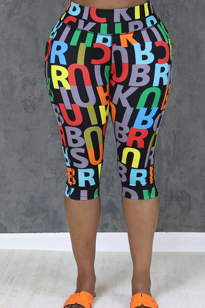 Only the lower half of a person is visible as they stand against a gray background, wearing Brick Built’s Lingo Pop Capris—high waist, knee-length capris featuring colorful overlapping bold letters—and orange slide sandals.