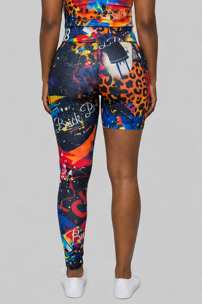 A person stands facing away, wearing the Art Code One-Legged Tights in multi—a matching colorful outfit with one shorts leg and one full leggings leg, crafted from polyester spandex with abstract patterns and text—and white sneakers, set against a plain gray background.