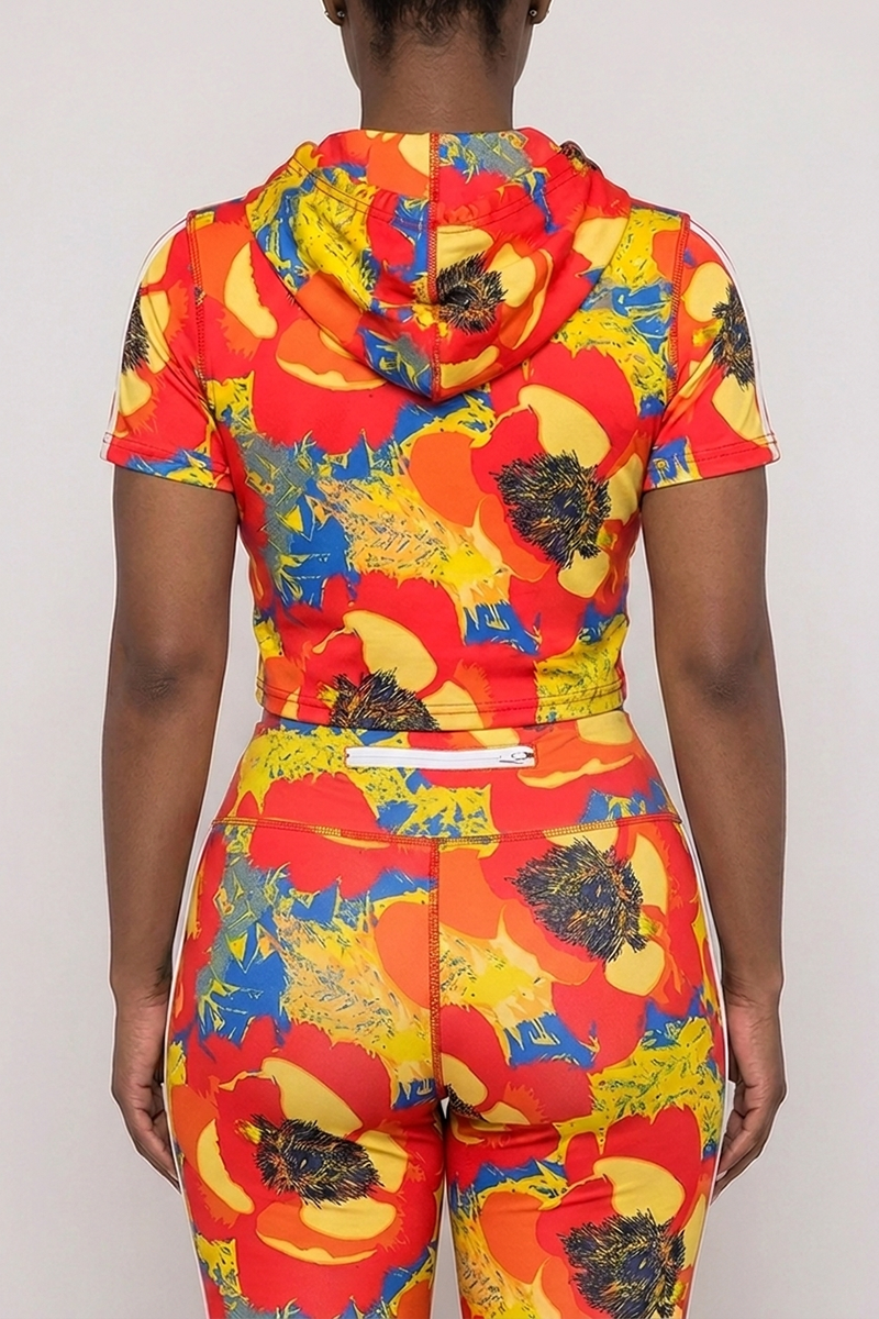 A person is shown from the back, wearing the Orange Crush Hooded Top with short sleeves and drawstrings, paired with matching pants featuring a bold red, yellow, and blue floral pattern set against a neutral background. The outfit is crafted from a comfortable polyester spandex blend for ease of movement.