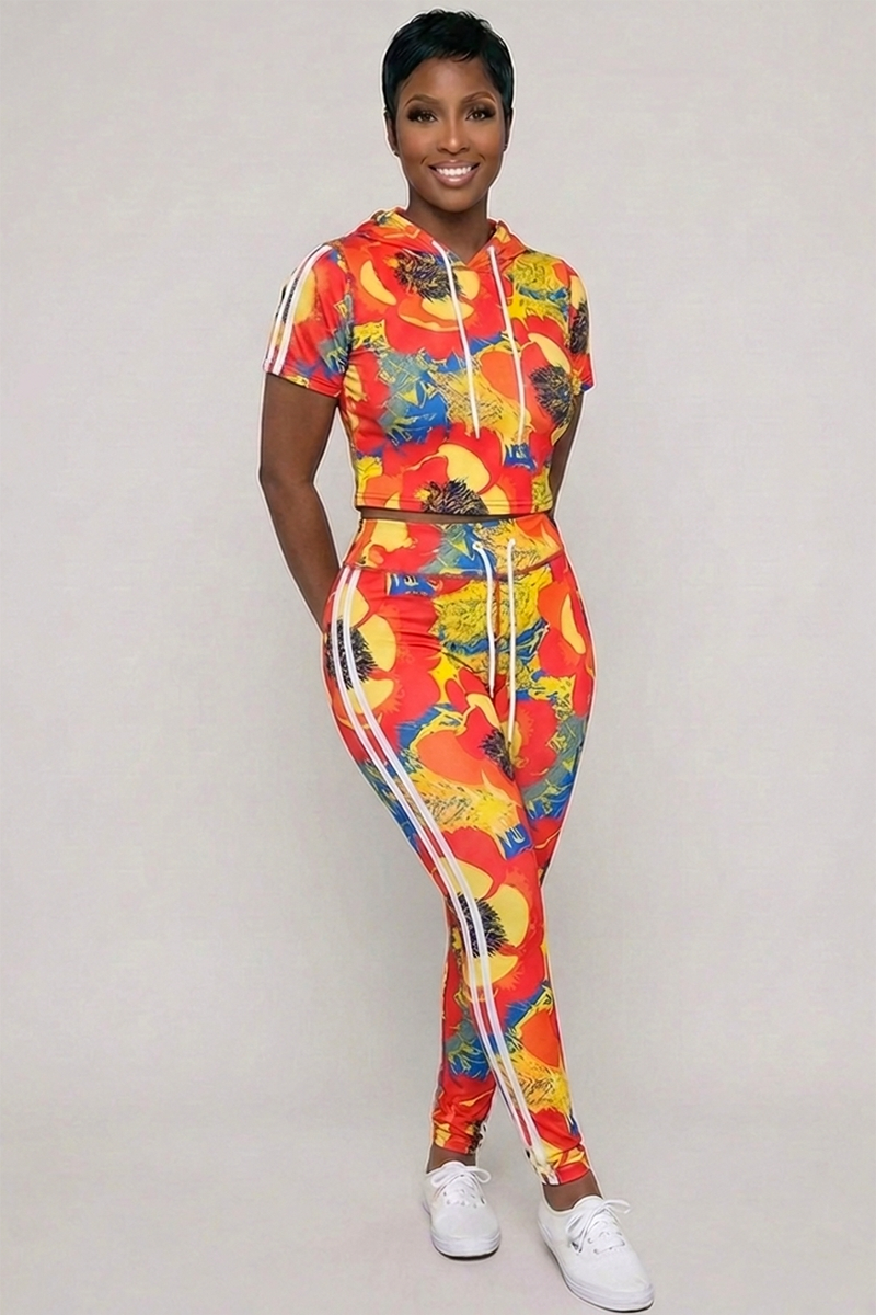 A woman stands against a plain background, wearing the Orange Crush Tights—a colorful, body-contouring ensemble featuring a fitted short-sleeve hoodie and matching pants in vivid red, yellow, and blue. The outfit flatters her silhouette and is paired with white sneakers. She smiles with her hands behind her back.