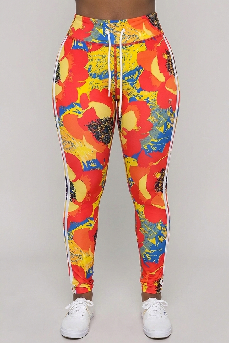 A person stands facing forward, wearing Orange Crush Tights—colorful floral 4-way stretch leggings with red, yellow, and blue patterns—paired with white sneakers. The leggings feature a body-contouring fit, white drawstring, and side stripes. The background is plain light gray.