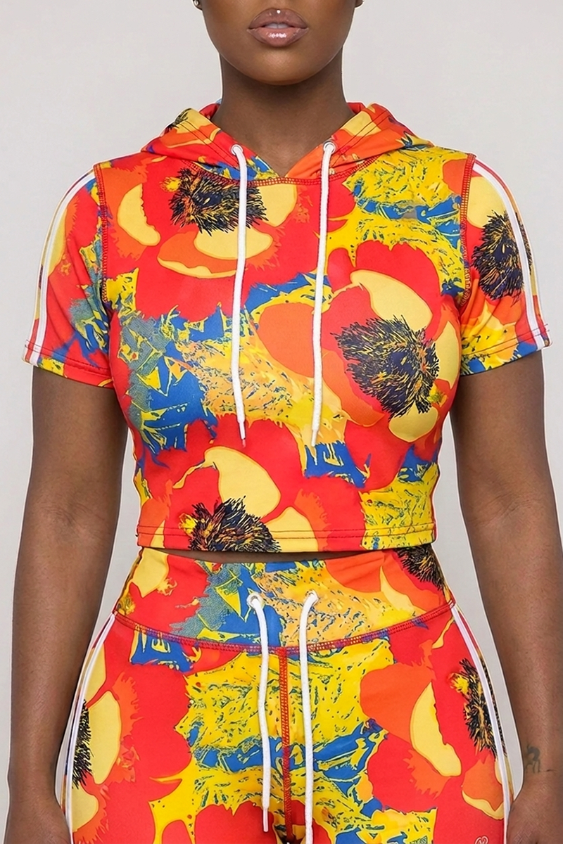 A person wears the Orange Crush Hooded Top, a short-sleeve, hooded crop top with white drawstrings, paired with matching pants. The multi-colored floral pattern in red, yellow, and blue stands out against the soft polyester spandex fabric, which offers comfortable 4-way stretch. The scene is set against a plain light gray background.