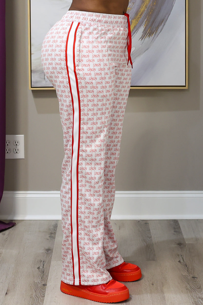 A person is indoors wearing BB Monogram Sweatpants in red-cream with a repeating pattern, red side stripes, zipper pockets, an elastic drawstring waistband, and bright red shoes. A painting and electrical outlet are in the background.