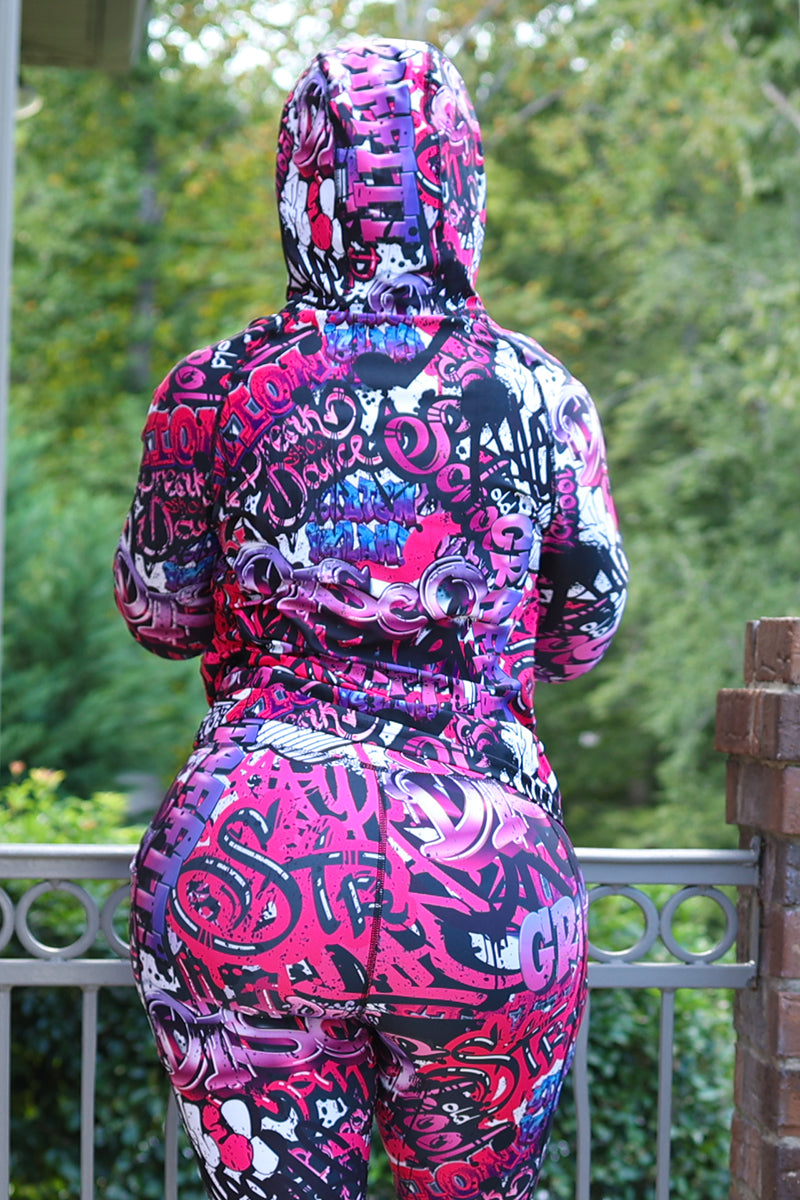 A woman wearing a Pink Graffiti Pullover, featuring a unique graffiti design, stretchy fabric, and a drawstring hood, perfect for workouts or casual outings.