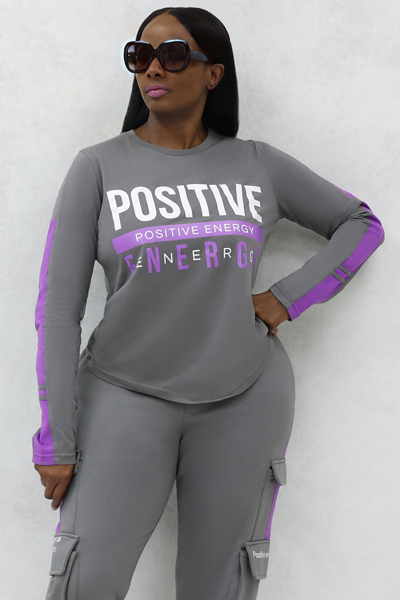 A woman wearing black sunglasses in a gray and purple tracksuit with the word "POSITIVE ENERGY" printed on it.