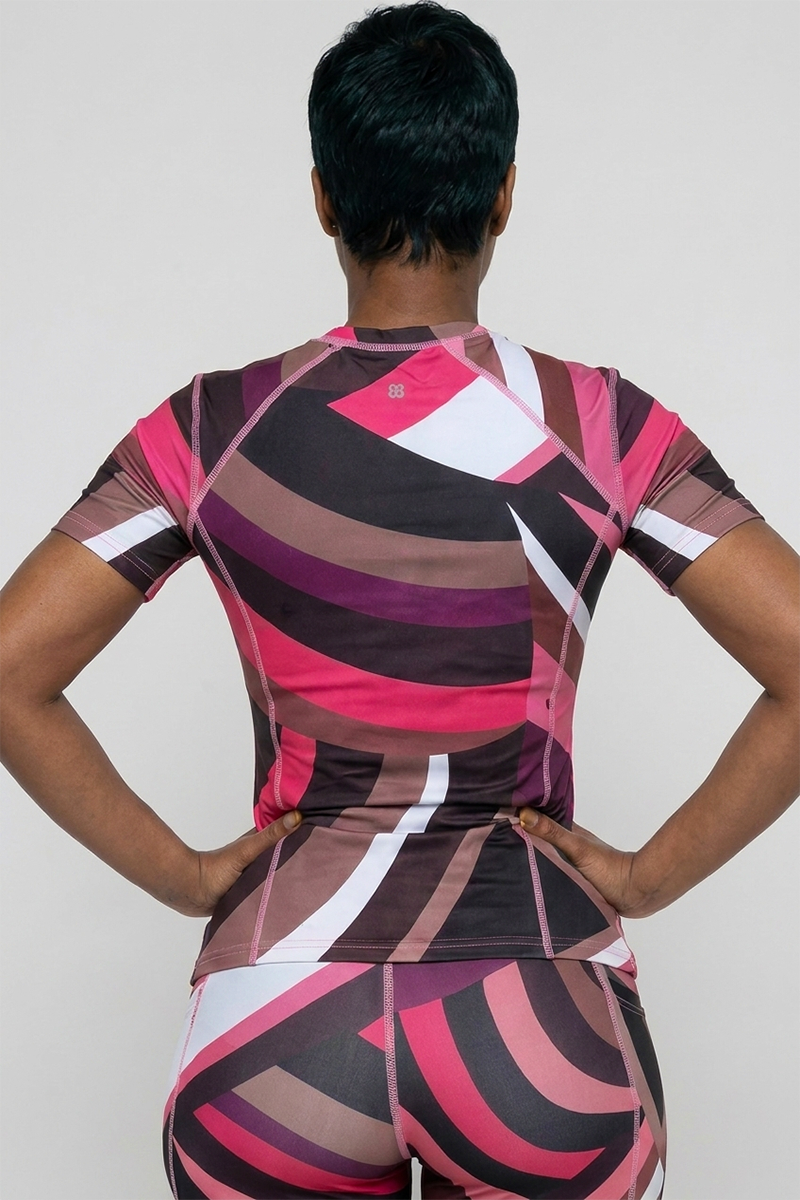 A person with short dark hair stands with hands on hips, facing away, wearing the Sable Tshirt—an activewear top in a geometric pattern, made from stretchy, lightweight, breathable fabric in pink, brown, black, and white.