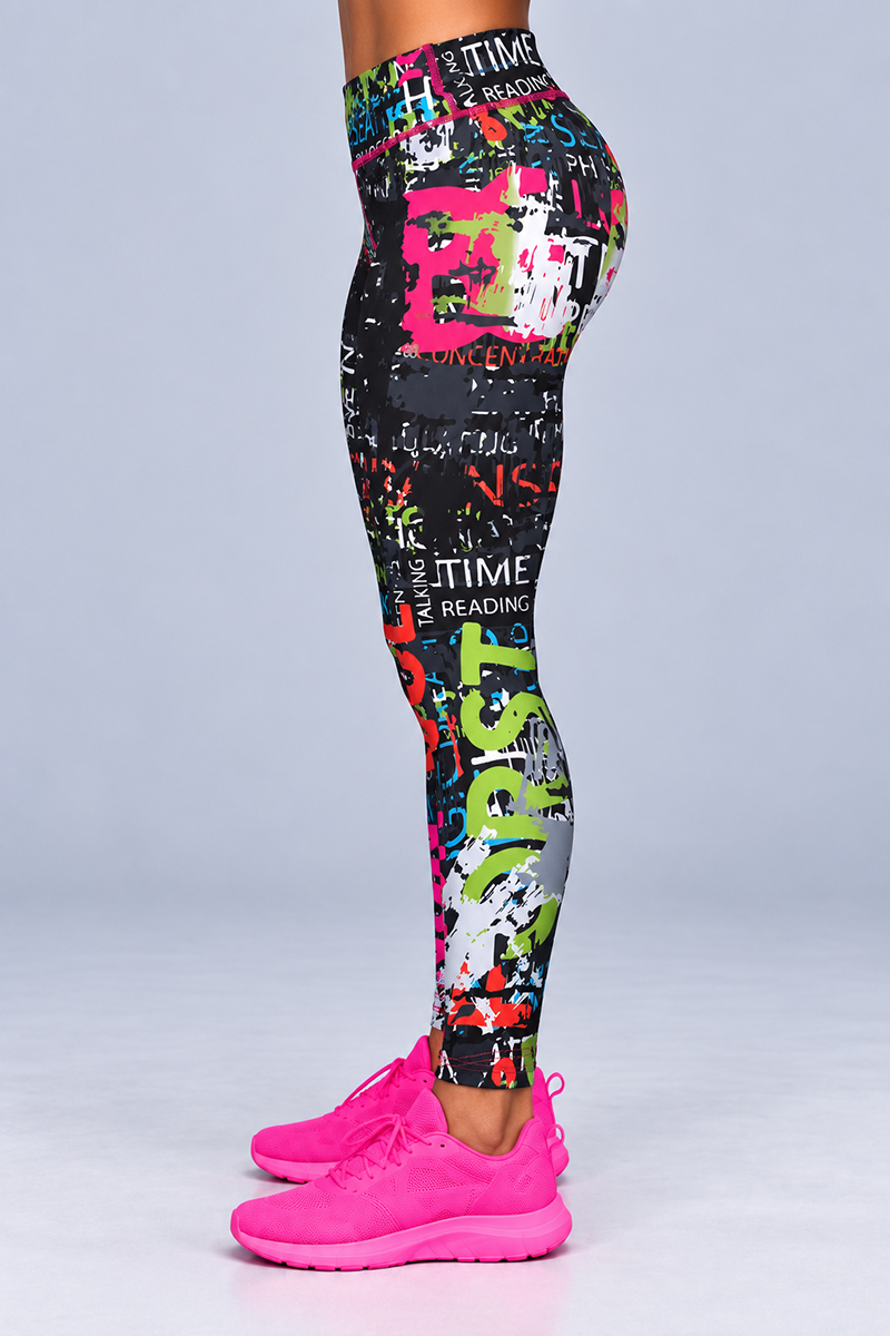 A person stands sideways against a plain light grey background, wearing Electric Letters Tights—colorful, graffiti-style stretchy leggings featuring various words—paired with pink athletic shoes. The body-contouring fit of the tights highlights the silhouette while providing both comfort and style.