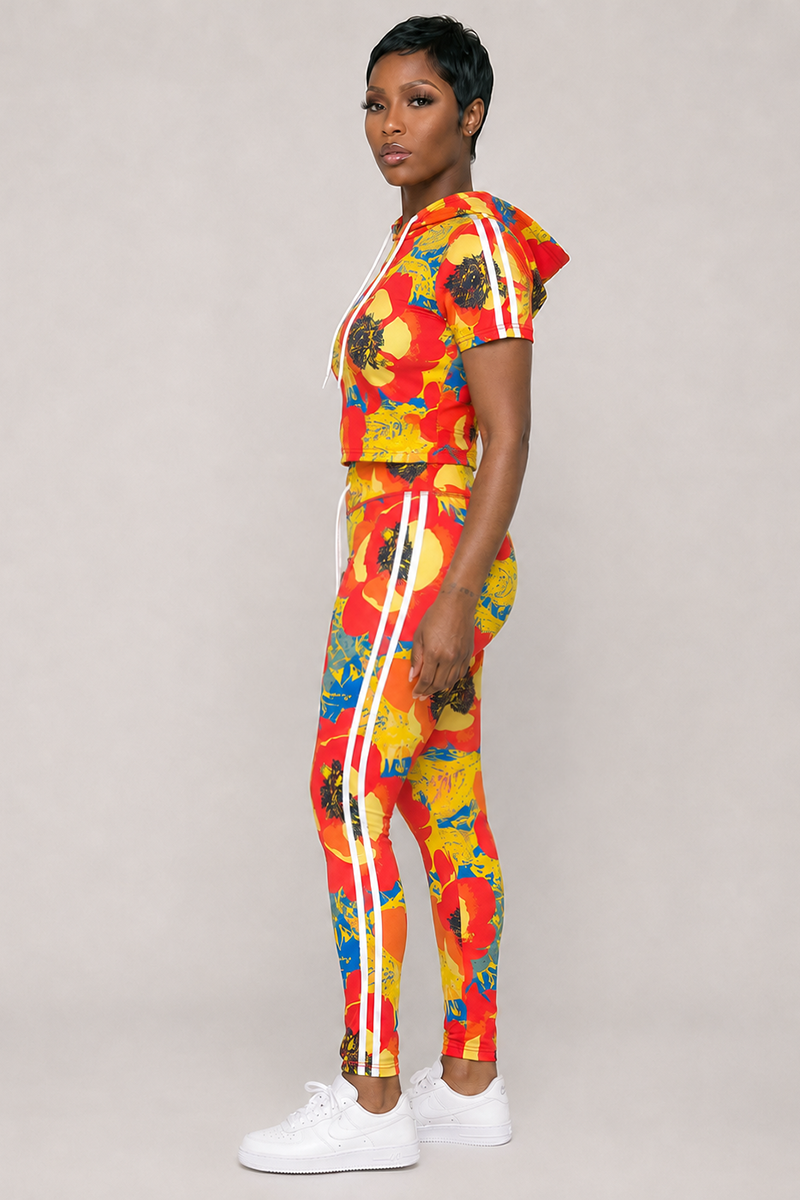 A person stands sideways wearing the Orange Crush Hooded Top—a vibrant red, yellow, and blue floral print tracksuit with white stripes and a hooded drawstring, crafted from a comfortable polyester spandex blend for 4-way stretch—paired with white sneakers against a plain light gray background.