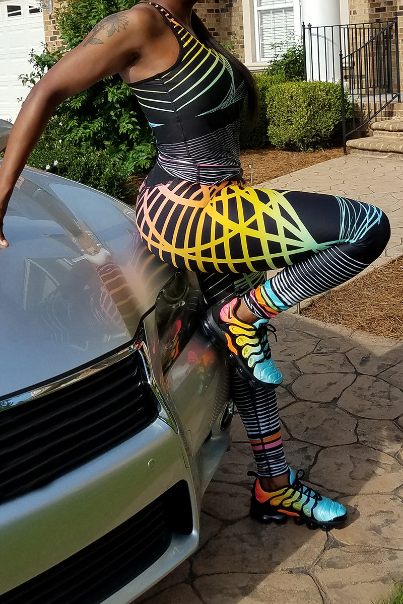Wearing Street Gradient Tights and matching sneakers, a person (head out of frame) leans against a silver car in a driveway, surrounded by greenery and a house in the background. The tights feature colorful geometric patterns and are anti-chafing workout wear.