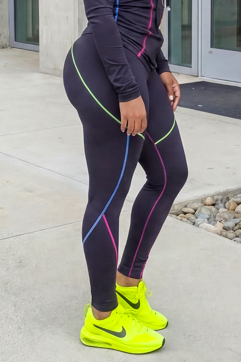 A person stands outside a building entrance, wearing Multi-Stitching Tights featuring colorful lines and a matching top with bright yellow Nike sneakers. The stretchy tights offer both comfort and style. Their face is not visible in the image.
