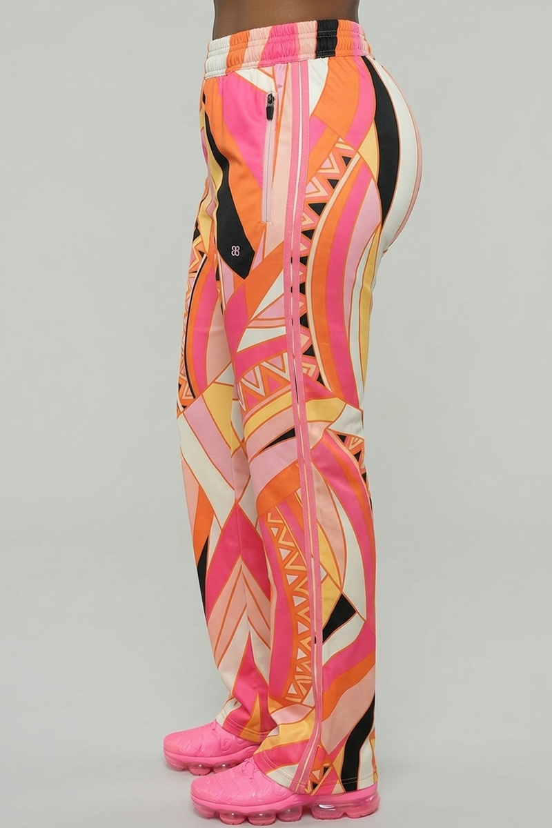 A person wears Vanta Pants—high-waisted, wide-leg, stretchy pants with a vibrant pink, orange, and yellow geometric pattern—paired with bright pink sneakers. The image is a side view against a plain background.