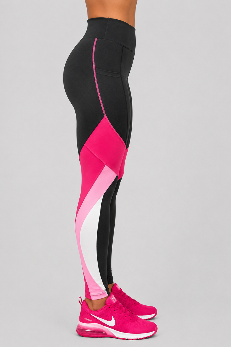 A person stands in profile against a plain light gray background, wearing the Velocity Tights—black leggings with pink and white color block accents designed for a body-contouring fit that creates a flattering silhouette—paired with bright pink sneakers.