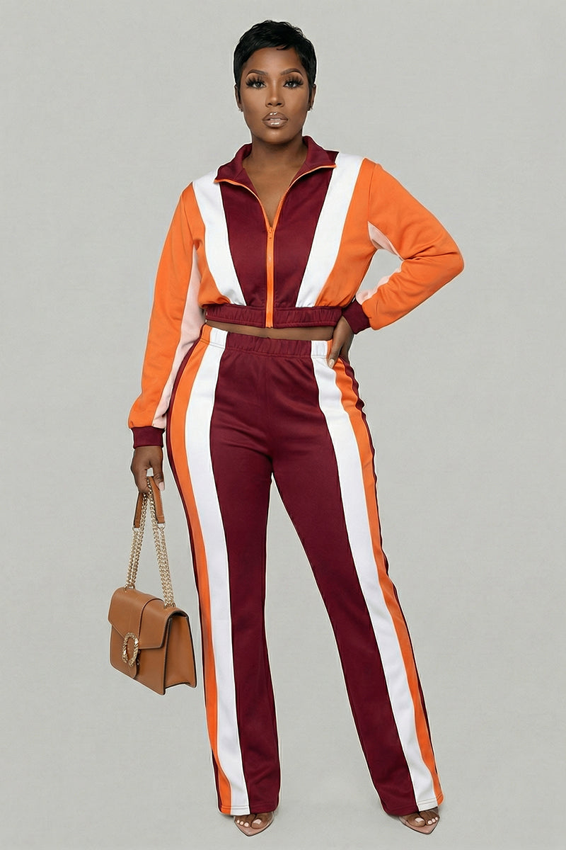 A woman with short hair stands against a plain background wearing Color Block SweatPants in burgundy, orange, and cream with an elastic waistband. She holds a tan handbag.